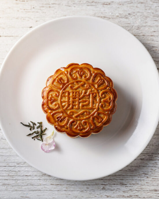 A golden-brown mooncake sits on a rustic wooden board, sliced to reveal a center of deep red bean paste.