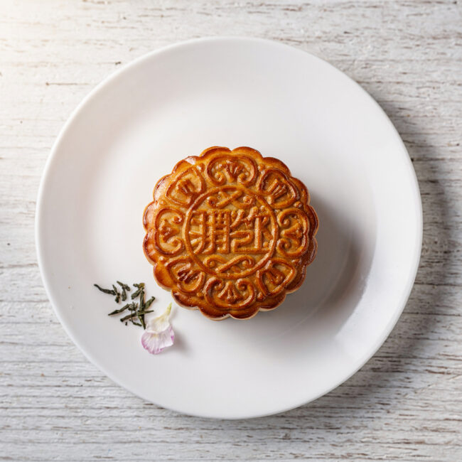 A golden-brown mooncake sits on a rustic wooden board, sliced to reveal a center of deep red bean paste.