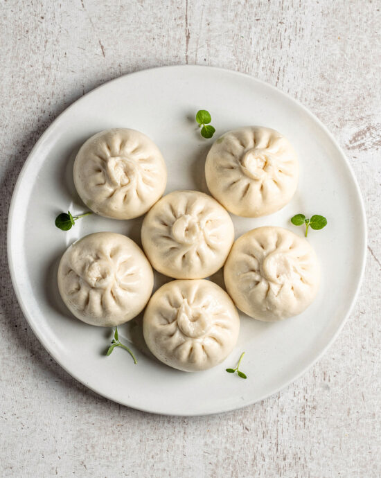 A steaming bamboo basket filled with soft, white bao buns on a rustic wooden table.