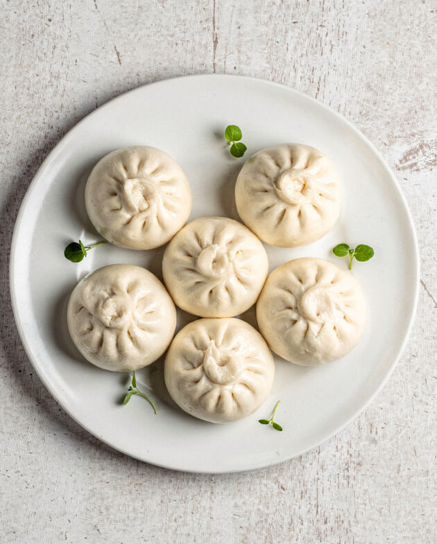 A steaming bamboo basket filled with soft, white bao buns on a rustic wooden table.