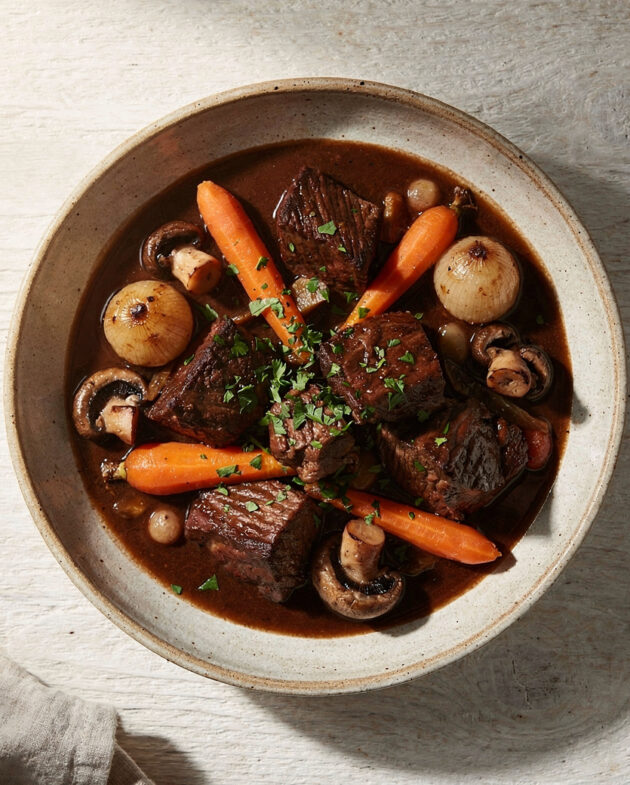 A steaming bowl of the finished beef bourguignon recipe, garnished with parsley, sits on a rustic wooden table.