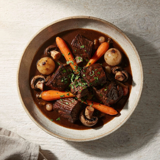 A steaming bowl of the finished beef bourguignon recipe, garnished with parsley, sits on a rustic wooden table.