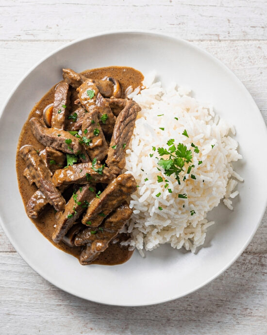 A creamy and inviting bowl of beef stroganoff brazil served with white rice and crispy potato sticks.