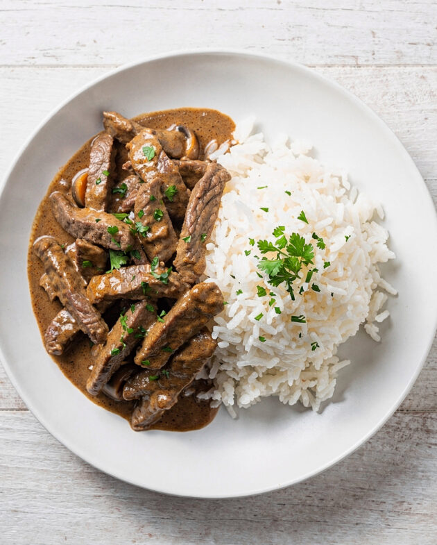 A creamy and inviting bowl of beef stroganoff brazil served with white rice and crispy potato sticks.