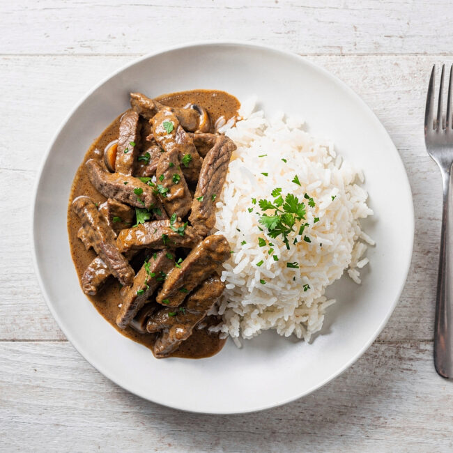 A creamy and inviting bowl of beef stroganoff brazil served with white rice and crispy potato sticks.