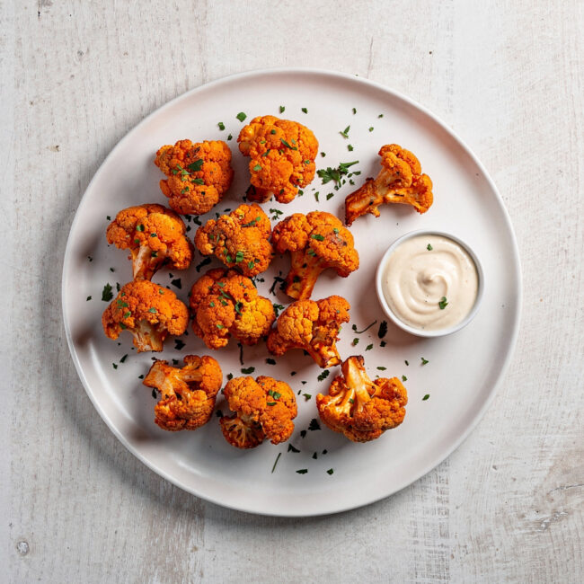 A golden-brown batch of crispy buffalo cauliflower served with creamy dip and celery sticks on a rustic wooden board.