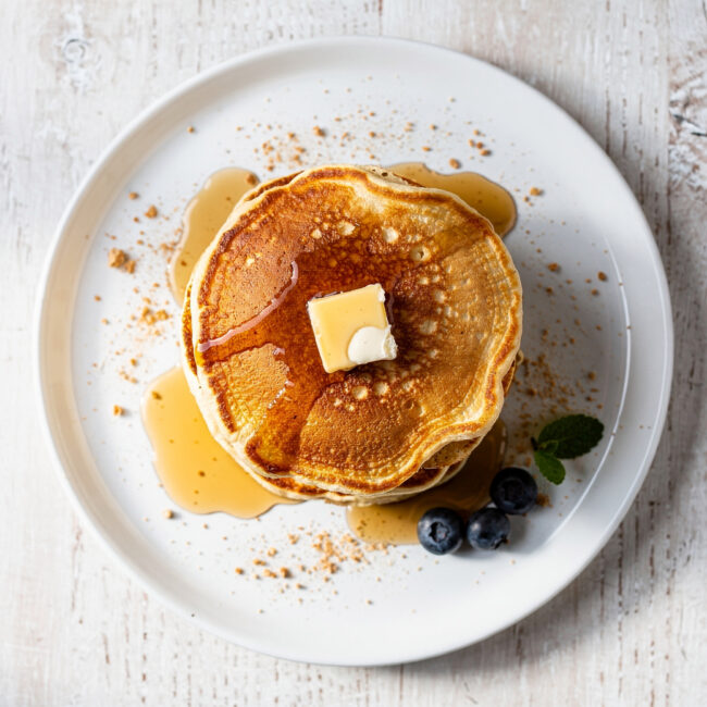 A golden stack of perfect buttermilk pancakes from our favorite buttermilk pancake recipe, dripping with maple syrup.
