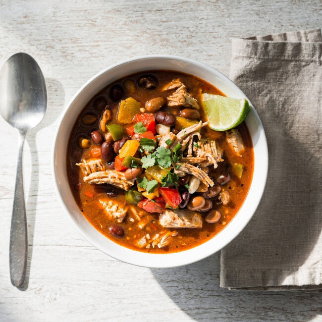 A vibrant bowl of this easy chicken chili recipe, topped with avocado and cilantro, sits on a rustic wooden table.