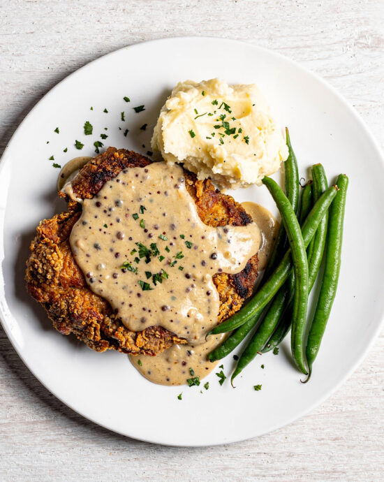 A perfectly golden piece of chicken fried steak recipe sits atop creamy gravy with a side of mashed potatoes.