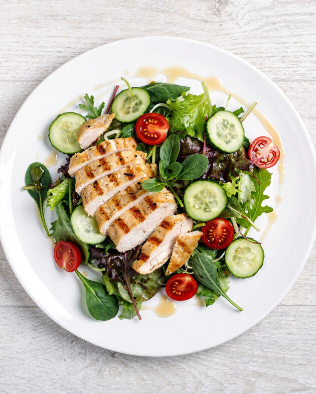 A creamy chicken salad with chunks of chicken, celery, and grapes sits in a white bowl on a wooden table.