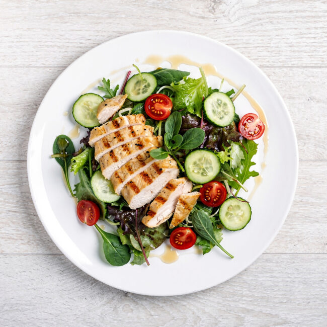 A creamy chicken salad with chunks of chicken, celery, and grapes sits in a white bowl on a wooden table.