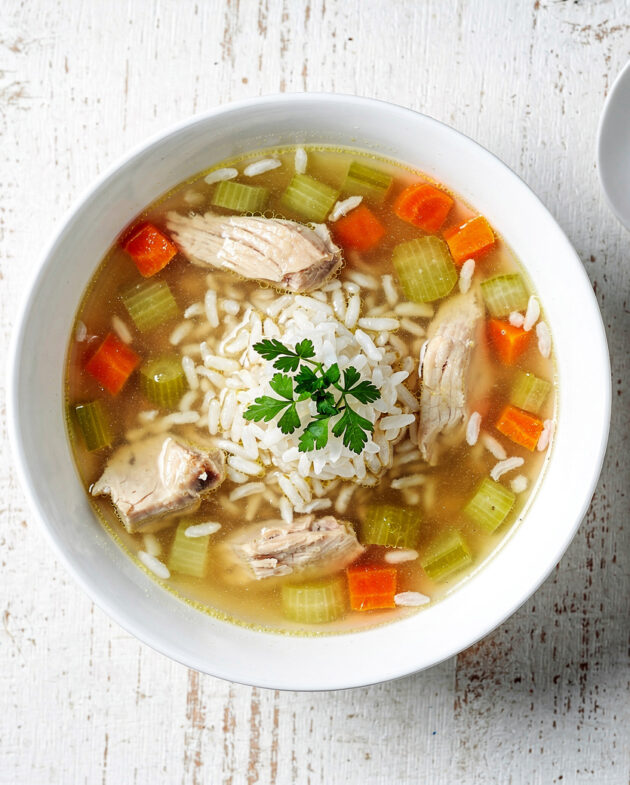 A steaming bowl of golden chicken soup with rice, carrots, and celery, photographed on a rustic wooden table.