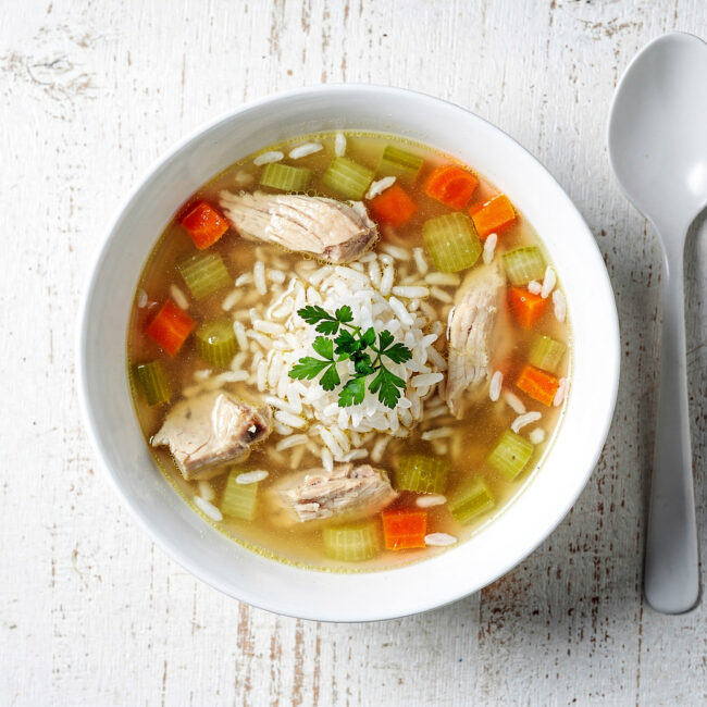 A steaming bowl of golden chicken soup with rice, carrots, and celery, photographed on a rustic wooden table.