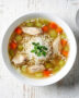 A steaming bowl of golden chicken soup with rice, carrots, and celery, photographed on a rustic wooden table.