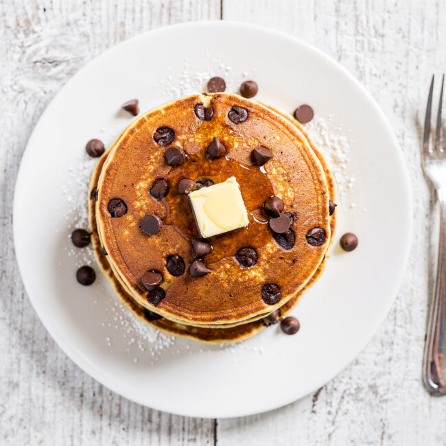 A tall stack of golden chocolate chip pancakes drizzled with syrup, ready for a cozy breakfast.