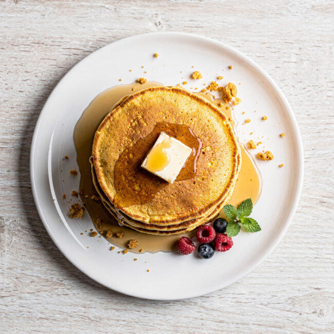 A stack of golden brown cornmeal pancakes drizzled with maple syrup on a rustic wooden board.