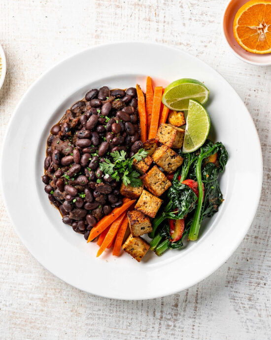 A steaming bowl of feijoada vegetarian with black beans, mushrooms, and plantains, served over rice with orange wedges.