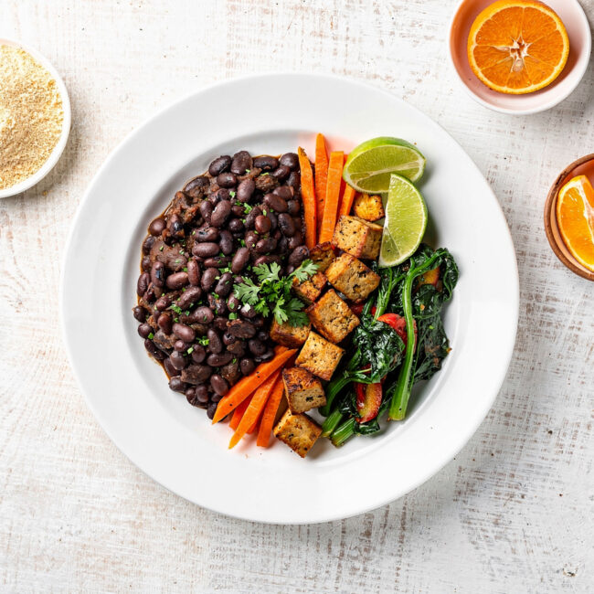 A steaming bowl of feijoada vegetarian with black beans, mushrooms, and plantains, served over rice with orange wedges.