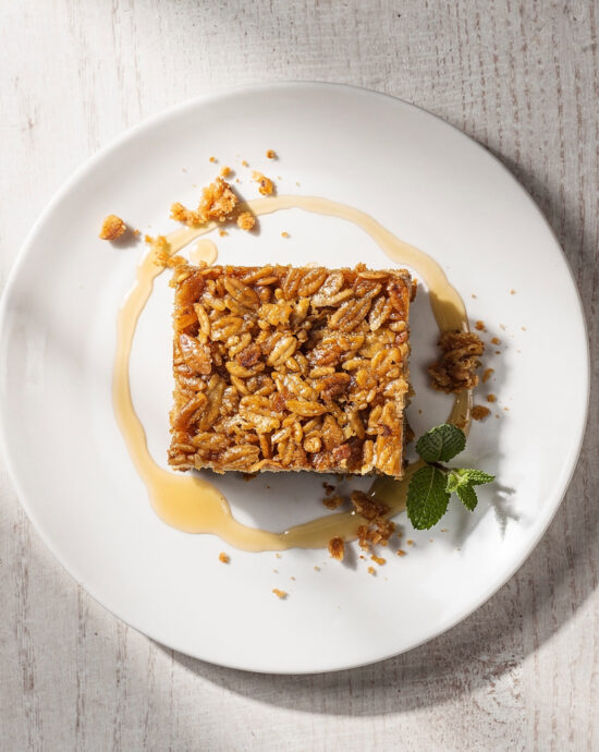 A golden-brown slab of chewy flapjack recipe, cut into squares and stacked, sits on a rustic wooden board.