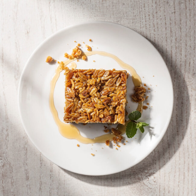 A golden-brown slab of chewy flapjack recipe, cut into squares and stacked, sits on a rustic wooden board.