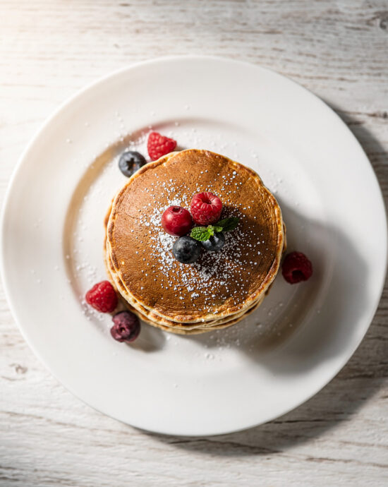 A tall, fluffy stack of golden-brown gluten free pancakes drizzled with maple syrup, shot with soft natural light.