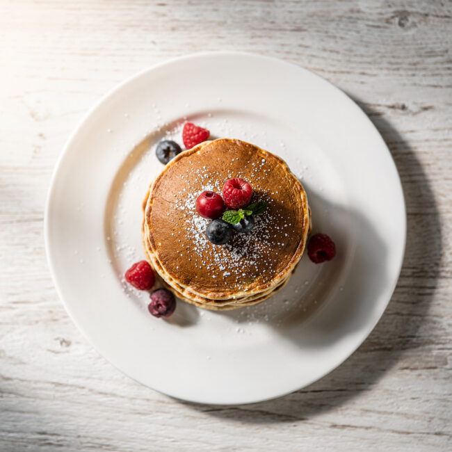 A tall, fluffy stack of golden-brown gluten free pancakes drizzled with maple syrup, shot with soft natural light.