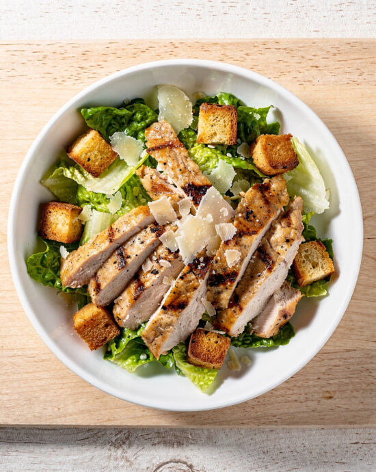 A beautiful, fresh grilled chicken caesar salad plated on a rustic wooden board, featuring smoky chicken and a creamy dressing.