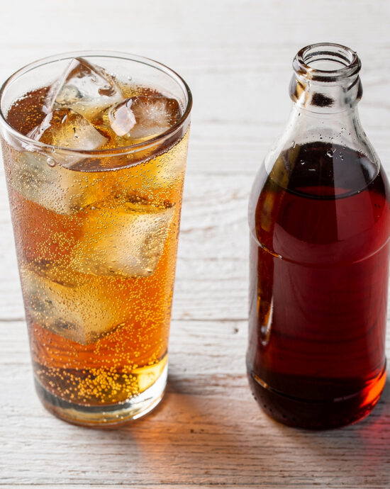 A tall, frosty glass of homemade guarana soda with ice and a slice of lime, beading with condensation on a rustic wooden table.