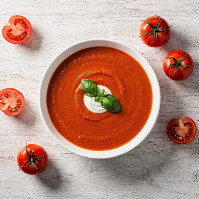 A vibrant bowl of homemade tomato soup with fresh tomatoes, garnished with basil and a swirl of cream, sits on a rustic wooden table.