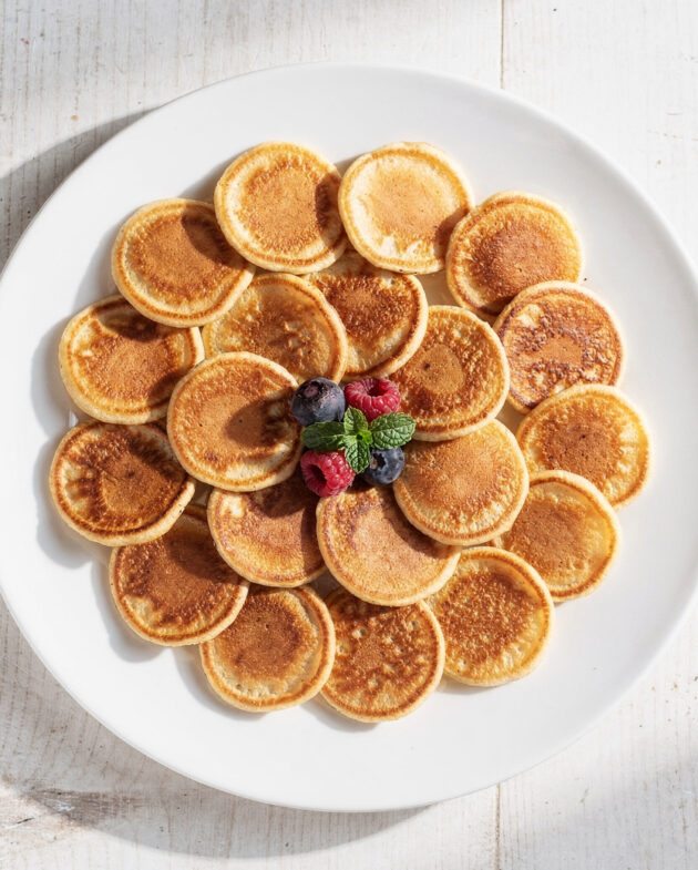 A stack of golden mini pancakes drizzled with maple syrup on a wooden board.