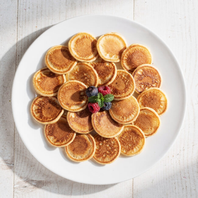 A stack of golden mini pancakes drizzled with maple syrup on a wooden board.