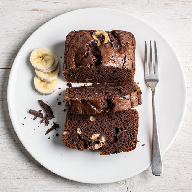 A perfectly baked slice of moist chocolate banana loaf recipe sits on a rustic wood board.