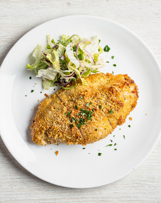 A golden and crispy parmesan crusted chicken breast plated elegantly with lemon wedges and fresh herbs.