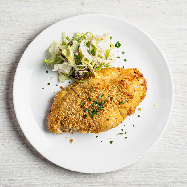 A golden and crispy parmesan crusted chicken breast plated elegantly with lemon wedges and fresh herbs.
