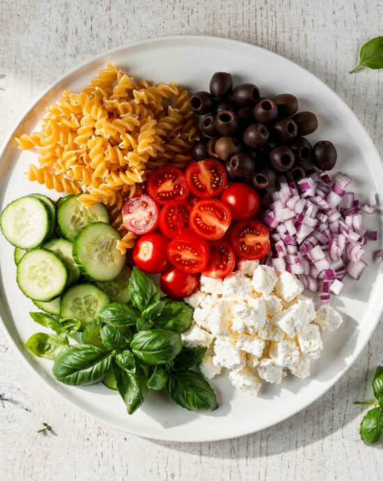 An overhead view of vibrant pasta salad ingredients neatly arranged on a wooden table.