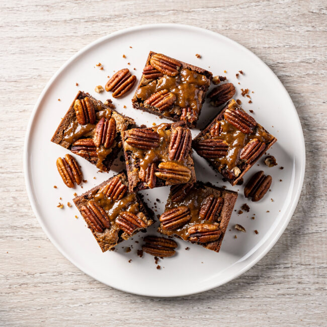 A golden brown pecan topping glistens over a dark, fudgy brownie base in a pan of freshly baked pecan pie brownies.