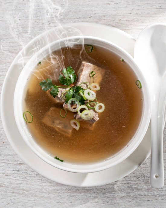 A rich, amber-hued pork bone broth served in a simple white bowl, garnished with fresh chives.