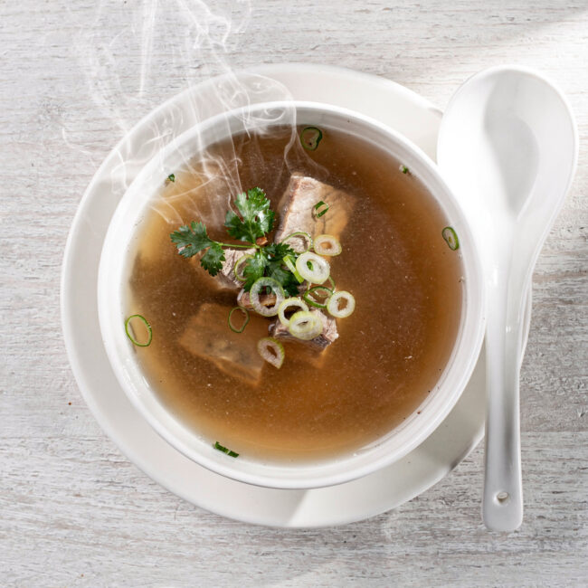 A rich, amber-hued pork bone broth served in a simple white bowl, garnished with fresh chives.