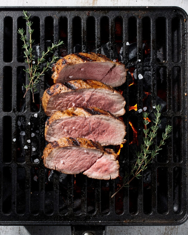 A beautifully grilled pork tenderloin on charcoal grill, sliced and served on a wooden platter.