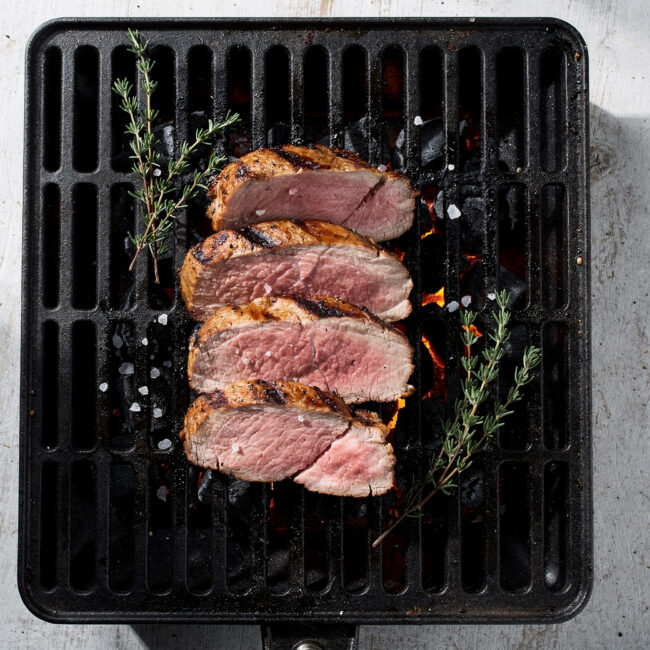 A beautifully grilled pork tenderloin on charcoal grill, sliced and served on a wooden platter.