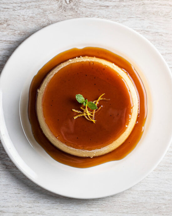 A perfect, golden-brown slice of pudim de leite sitting in a pool of dark caramel sauce.