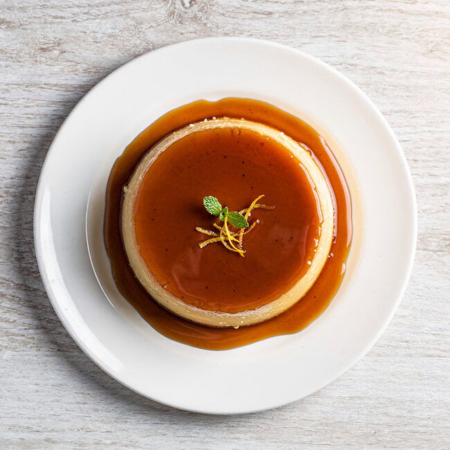 A perfect, golden-brown slice of pudim de leite sitting in a pool of dark caramel sauce.