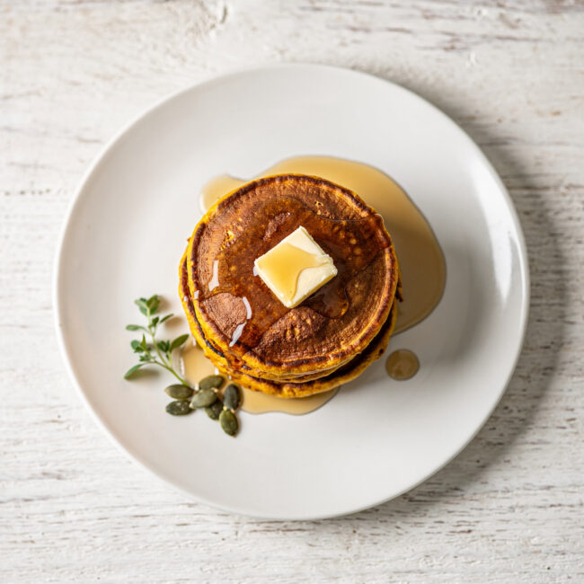 A golden stack of fluffy pumpkin pancake recipe drizzled with maple syrup on a rustic wooden table.