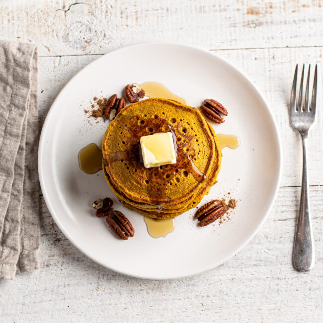 A golden stack of fluffy pumpkin pancakes from this classic pumpkin pancake recipe, drizzled with maple syrup.