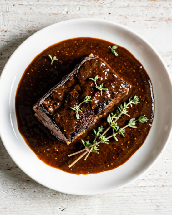 A glossy, dark short rib sauce being drizzled over tender braised beef in a rustic bowl.
