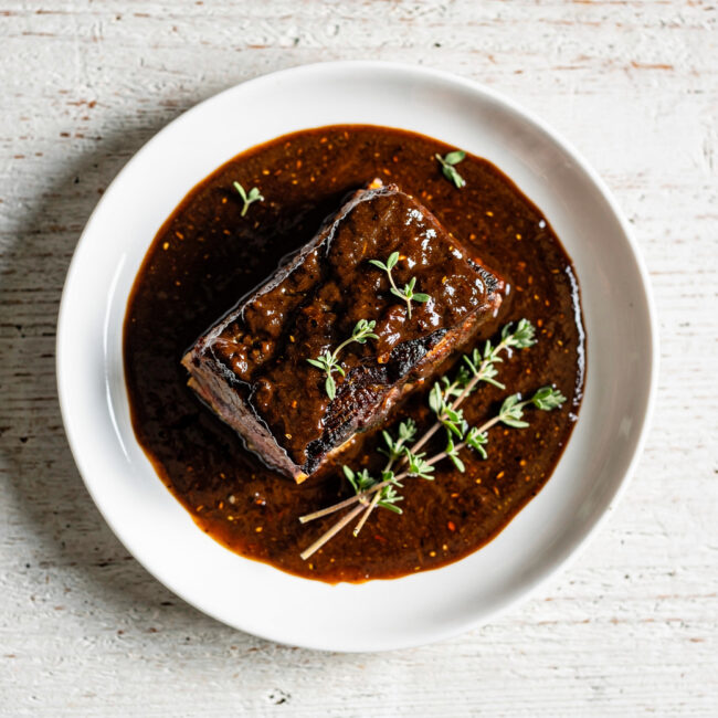 A glossy, dark short rib sauce being drizzled over tender braised beef in a rustic bowl.