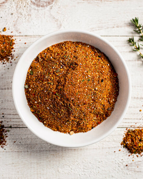 A jar of homemade spare ribs rub with a sprinkle on a wooden board next to a sprig of rosemary.