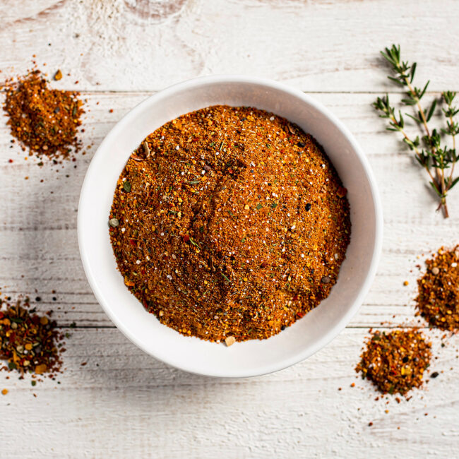 A jar of homemade spare ribs rub with a sprinkle on a wooden board next to a sprig of rosemary.