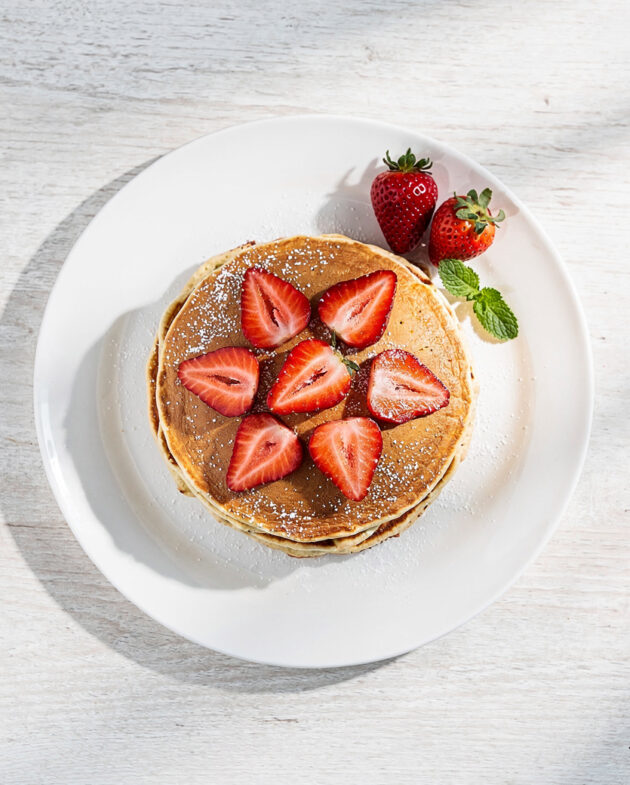 A tall stack of golden, fluffy strawberry pancakes drizzled with maple syrup on a rustic wooden table.