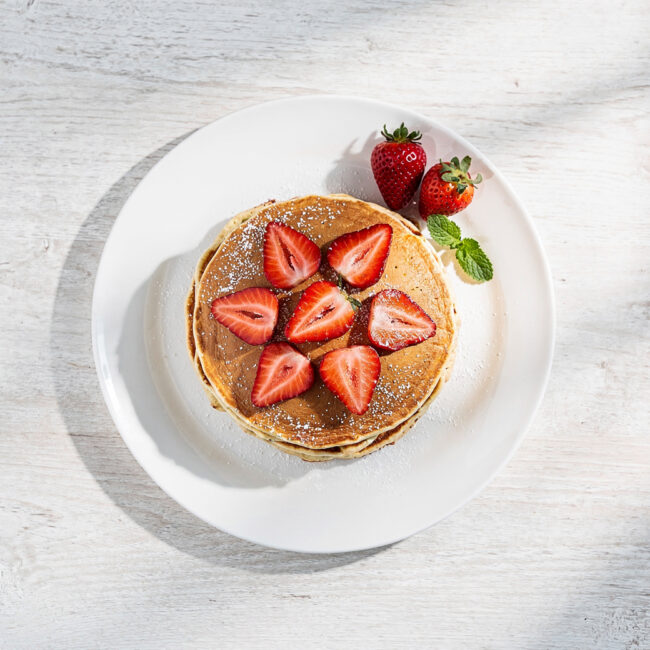 A tall stack of golden, fluffy strawberry pancakes drizzled with maple syrup on a rustic wooden table.